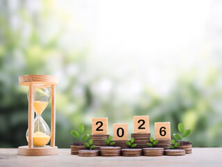 Hourglass, Wooden blocks with the number 2026 and plants growing up on stack of coins. The concept...