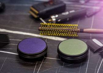 Two unbranded grooming product tins with colored lids on a barber workstation, surrounded by hair styling tools and accessories in a modern salon setting