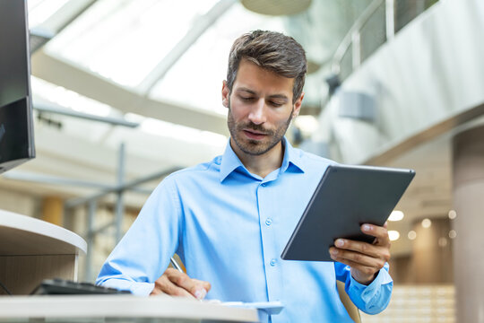 Smiling busy young employee, young entrepreneur using tablet computer, happy businessman executive looking at tab device analyzing finance trading market data working sitting at office desk.