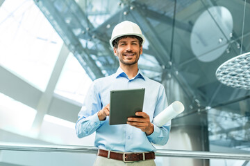 Handosme man with worker's helmet holding digital tablet while looking at camera in building under construction