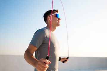 Man skipping rope on rooftop at sunset wearing sunglasses and earphones