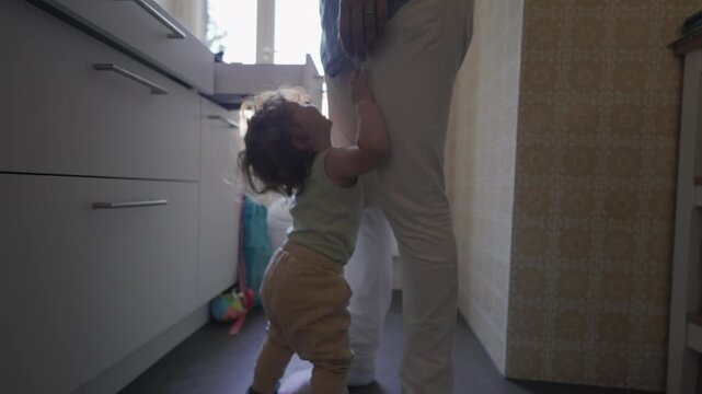 Toddler clinging to parent&rsquo;s leg in kitchen with arms raised and complaining expression, asking to be picked up while parent stands holding phone nearby
