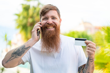 Redhead man with beard keeping a conversation with the mobile phone and holding a credit card