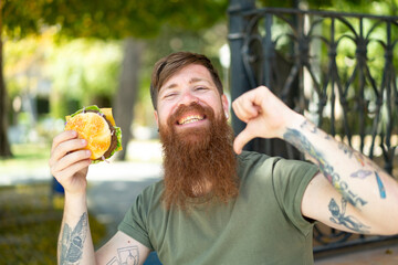 Redhead man with beard holding a burger at outdoors proud and self-satisfied
