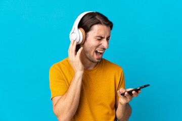Young handsome man isolated on blue background listening music with a mobile and singing