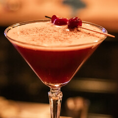 Close-up of a cherry cocktail in a martini glass with a foamy top and decorative garnish.