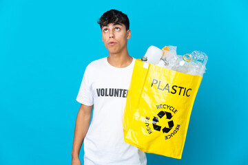 holding a bag full of plastic bottles to recycle over blue background looking up and with surprised...