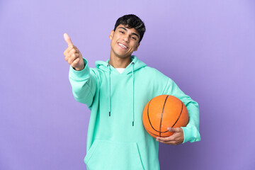 Young man playing basketball over isolated purple background with thumbs up because something good...