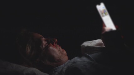 Older woman looking at smartphone in bed late at night, illuminated by screen light while scrolling through content in dark room - Powered by Adobe