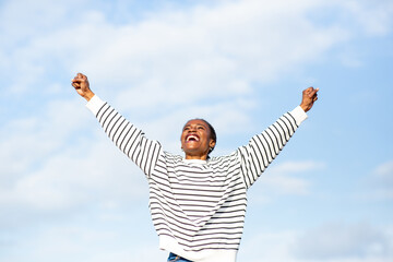 Excited African young woman raising arms in victory pose under blue sky