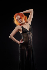 Fototapeta premium A woman in a black dress with red hair poses for the camera in a dark room. She has a beautiful figure. The girl is not wearing underwear under her dress.