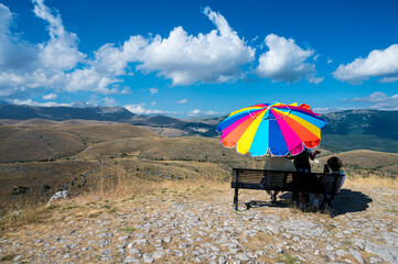 A happy family enjoys the mountain view with its blue sky and white clouds. They're in the shade of a huge, colorful umbrella. This image conveys a sense of peace and tranquility.