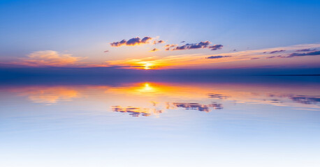 Obraz premium Brilliant hues of orange, purple, and blue paint the sky and reflect seamlessly on the tranquil water surface, capturing a serene and colorful evening view.