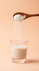 Wooden spoon pouring white powder into a glass of milk against a soft peach background