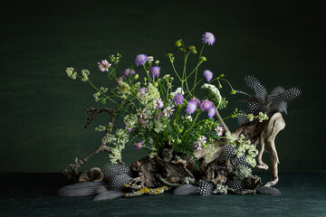 Composition of vibrant wildflowers, delicate guinea fowl feathers, lichens, and tree branches on a dark green background, radiating a mystical and mysterious atmosphere with expressive natural beauty.