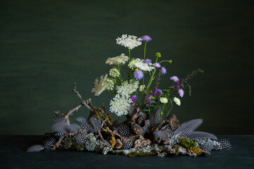 Composition of vibrant wildflowers, delicate guinea fowl feathers, lichens, and tree branches on a dark green background, radiating a mystical and mysterious atmosphere with expressive natural beauty.