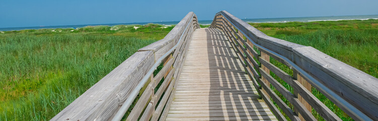 Panorama view upward view Boardwalk leads through coastal grass with shadows cast railings create a rhythmic pattern along planks, adding visual texture and depth toward Galveston beach, Texas