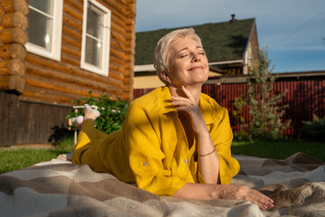 A serene scene of a mature woman dressed in yellow, leisurely relaxing on a blanket in her garden near house, enjoying sun