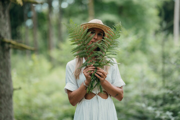 A woman’s face gently hidden behind forest fern, straw hat and blonde hair visible. Nature peace