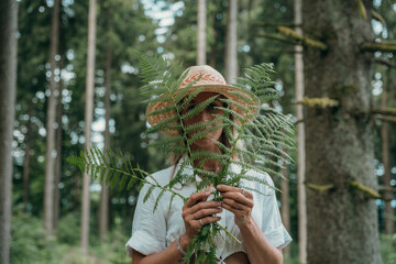 Blonde woman wearing straw hat behind green fern leaves in forest. Concept of nature, peace, and...