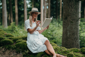 A woman sits on forest moss wearing a white dress and hat, reading peacefully