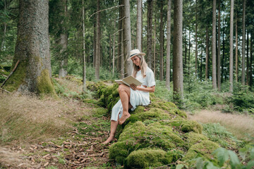 Reading in the forest, woman in dress and hat sits on moss in peaceful solitude