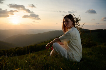 Woman sits peacefully on a grassy hill, gazing at the sunset as golden light bathes the landscape....