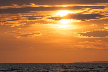 A vivid golden-orange sunrise lights up the sky above a calm sea, with clouds catching the warm glow and the coastline faintly visible in the distance.