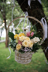 Basket of yellow and pink roses with ribbons gently sways from a tree branch. Surrounded by green...