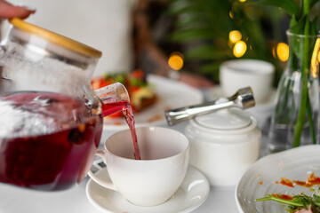 Close-up of a hand pouring red tea from a glass teapot into a white teacup on a saucer, with cozy tableware and plants in the background. Warm and inviting morning scene.