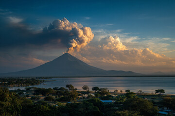Fototapeta premium A striking view of Momotombo volcano with smoke rising and Lake Managua glistening in the foreground at sunrise.