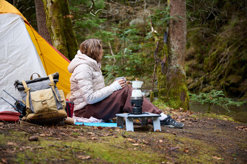 Hiker woman drinking coffee outdoor near the camping tent. A female traveler in the forest in early...