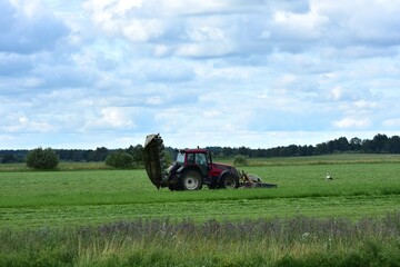 Red tractor working in a green field on a summer day. Rural farming equipment mowing or plowing grass in a flat agricultural landscape.