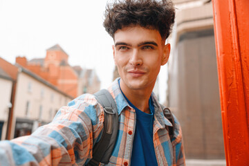 Young man takes a selfie in a vibrant city street during an overcast day while wearing a colorful...