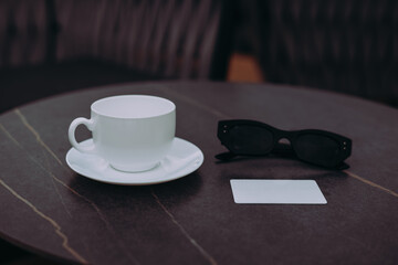 A white coffee cup, sunglasses, and card on a table. A white coffee cup with saucer, sunglasses, and blank card on a brown table, perfect for business and leisure.