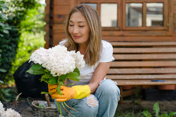 Woman with yellow gloves arranging fresh white hydrangeas in a garden near a wooden bench during a...