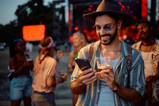 Happy festival goer drinking beer while using app on cell phone in front of music stage.