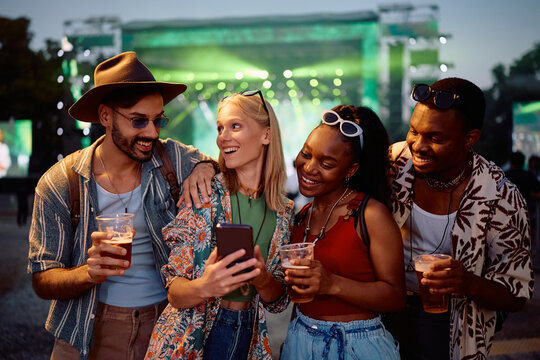 Multiracial group of happy festival goers using cell phone in front of music stage.
