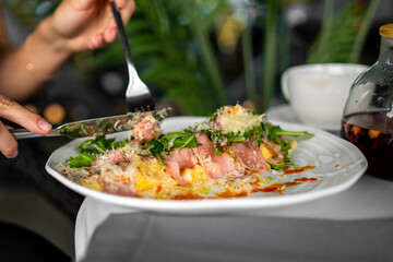 Close-up of a person eating a ham and cheese omelette with greens on a white plate, accompanied by a white cup and glass container on a breakfast table.