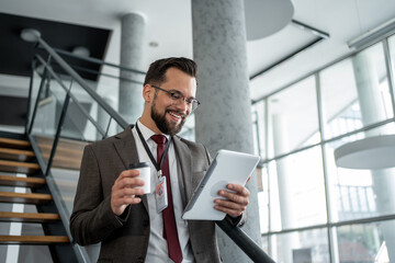 Fototapeta premium Happy businessman using digital tablet and drinking coffee on stairs in office building