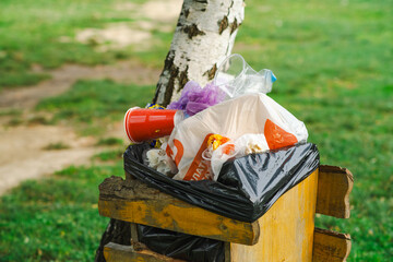 A garbage bin is overflowing with trash next to a tree in a park Brightly colored plastic waste and...
