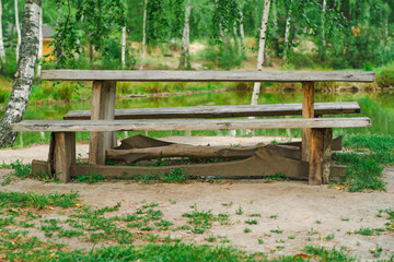 Naklejka premium A rustic wooden picnic table sits on sandy ground beside a peaceful body of water Towering trees and lush grass create a serene environment perfect for outdoor gatherings