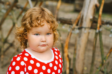 Child With Curly Hair in a Red Polka Dot Dress Stands Near Wooden Fence Outdoors in Natural Light...