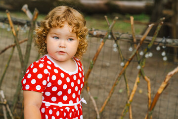 Child With Curly Hair in a Red Polka Dot Dress Stands Near Wooden Fence Outdoors in Natural Light...