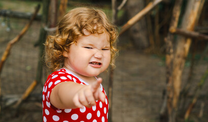 A young child with curly hair is expressing frustration by pointing and frowning in an outdoor...