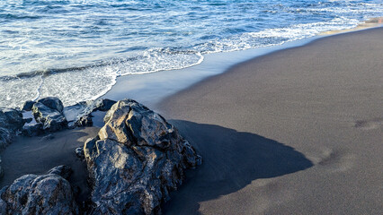 Surf waves close-up on a beach with black volcanic sand illuminated by the sun