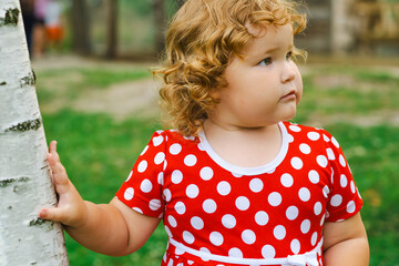 A young child with curly hair stands next to a birch tree in a vibrant green park The child is wearing a cheerful polka dot dress and is gazing thoughtfully into the distance surrounded by nature