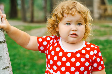 A young child with curly hair stands next to a birch tree in a vibrant green park The child is wearing a cheerful polka dot dress and is gazing thoughtfully into the distance surrounded by nature