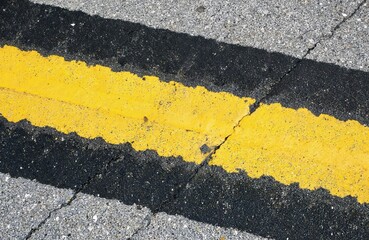 Close-up abstract view of asphalt road surface with yellow and black stripes. Textured pavement with cracks and worn paint offers geometric design for urban traffic and safety themes.