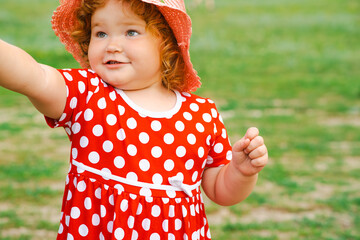 A young child wearing a bright red polka dot dress and a straw hat is happily reaching out with one hand in a green grassy area during a sunny day exuding joy and playfulness
